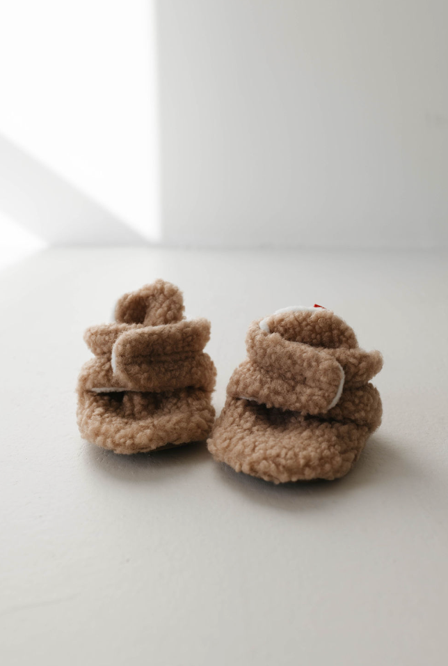 A pair of small, tan Sherpa Baby Booties by forever french baby, crafted with a soft, textured fabric and featuring adjustable elastic. The fuzzy booties are placed side by side on a light-colored surface against a bright white background.