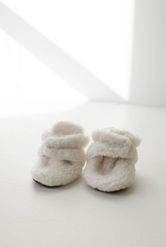 A pair of "Sherpa Baby Booties | Ivory" by forever french baby is placed on a white surface. The booties have a soft, textured appearance with adjustable elastic ankle straps, suggesting warmth and comfort for an infant. Bright light creates soft shadows, enhancing the cozy ambiance.