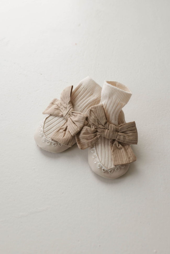 A pair of forever french baby's Ballerina Slipper Socks is displayed on a white surface, featuring attached moccasin-style soft sole shoes. The beige socks showcase vertical ribbing, and each shoe is embellished with an adorable big bow on the front, adding a cute and cozy touch.