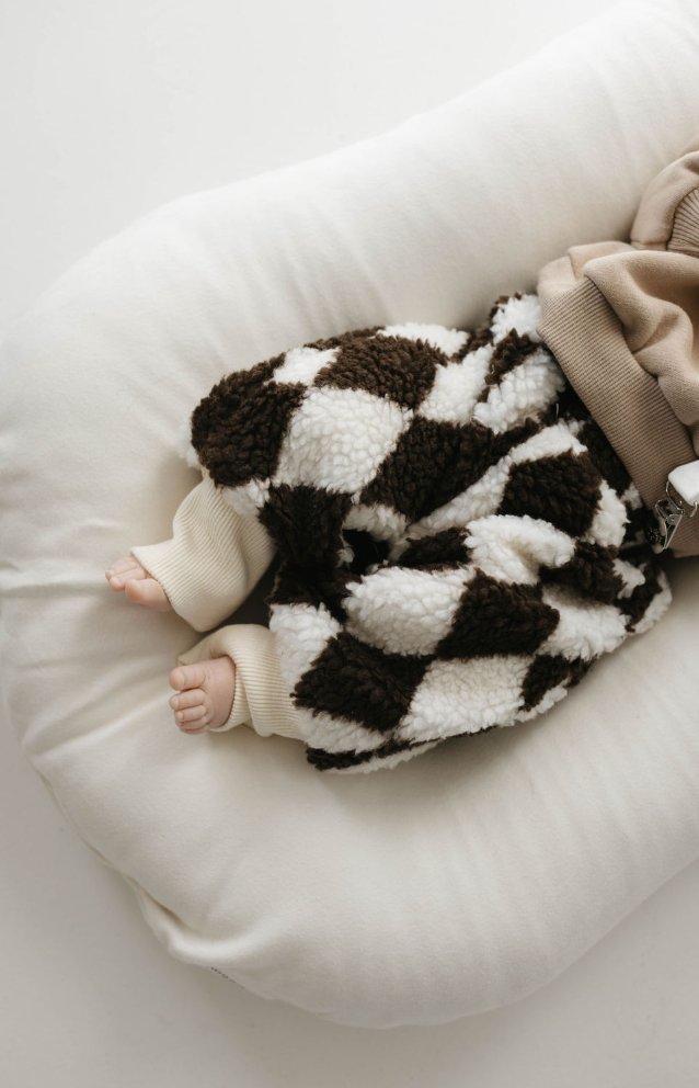 A baby lounges on a white cushion, wrapped in a cozy brown and white checkered blanket. Only the feet and part of a beige sleeve are visible, hinting at the Sherpa Brown Diamond Pants by Forever French Baby. The image captures winter comfort and fun beautifully.