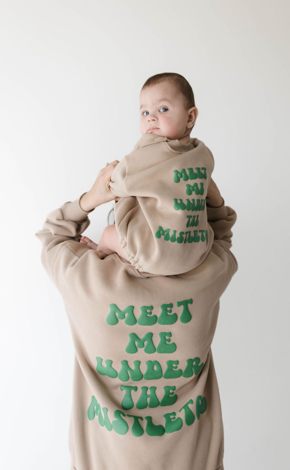 An adult and a baby are shown from the back, both wearing matching holiday sweatshirts in beige. The text on the back of these "Meet Me Under The Mistletoe" sweatshirts by forever french baby is written in green, puffy letters. The adult holds the baby up against a plain white background.