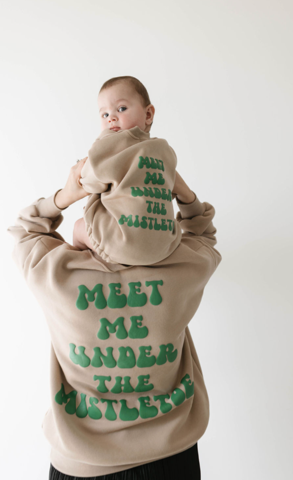 A person carries a baby on their shoulders, both celebrating the holiday spirit in matching beige attire by forever french baby. The adult's sweatshirt and the baby's long sleeve romper both read, "MEET ME UNDER THE MISTLETOE.