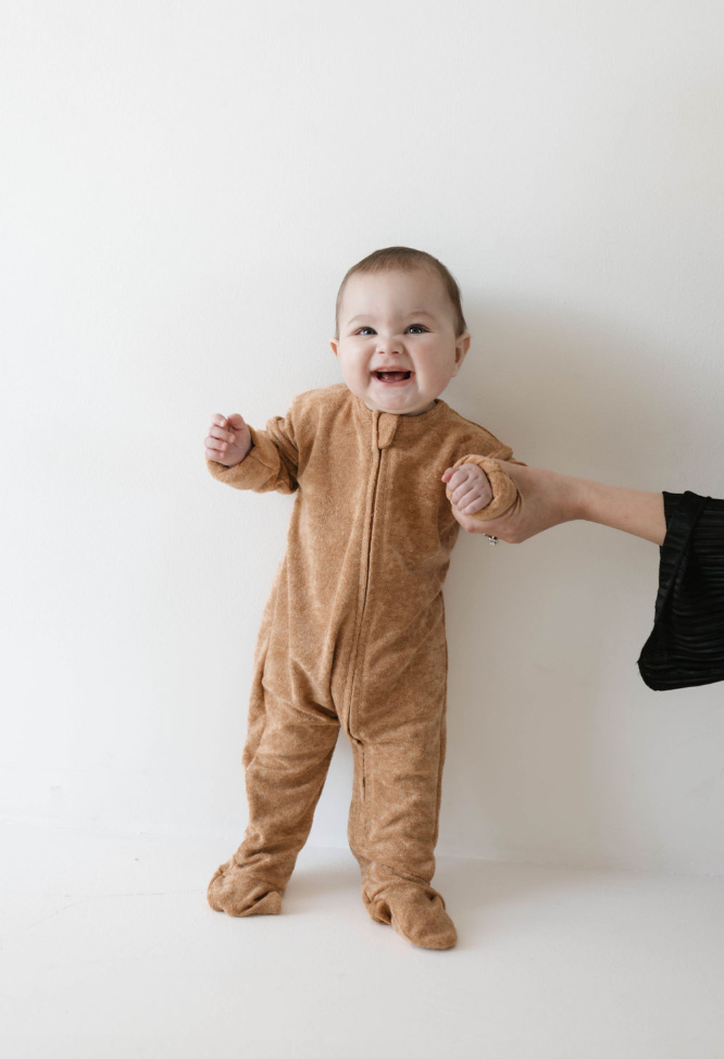 A baby, dressed in forever french baby's snug Brown Sugar | Terry Zip Pajamas, stands against a white background, smiling and clutching an adult's hand for support. The adult is partially visible, wearing a black long-sleeved top.