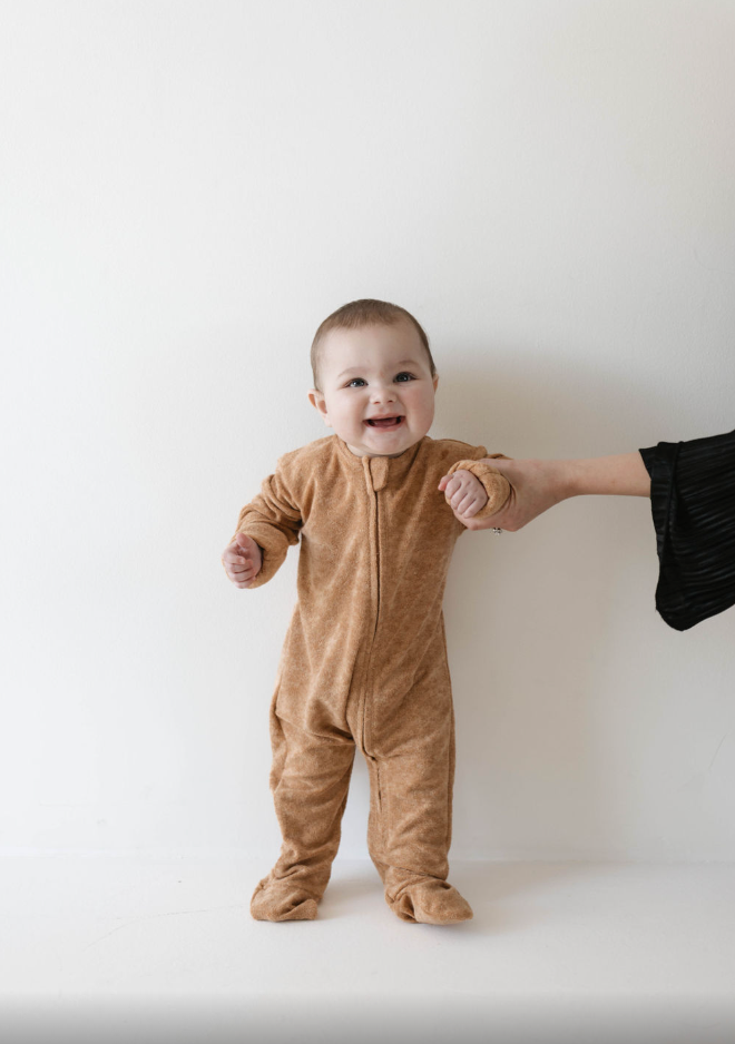 A smiling baby in a brown onesie stands while holding an adult's hand, who is partially visible on the right of the image. The background is a plain, light-colored wall, giving a cozy snuggle feel much like the Terry Zip Pajamas from forever french baby’s Brown Sugar collection.