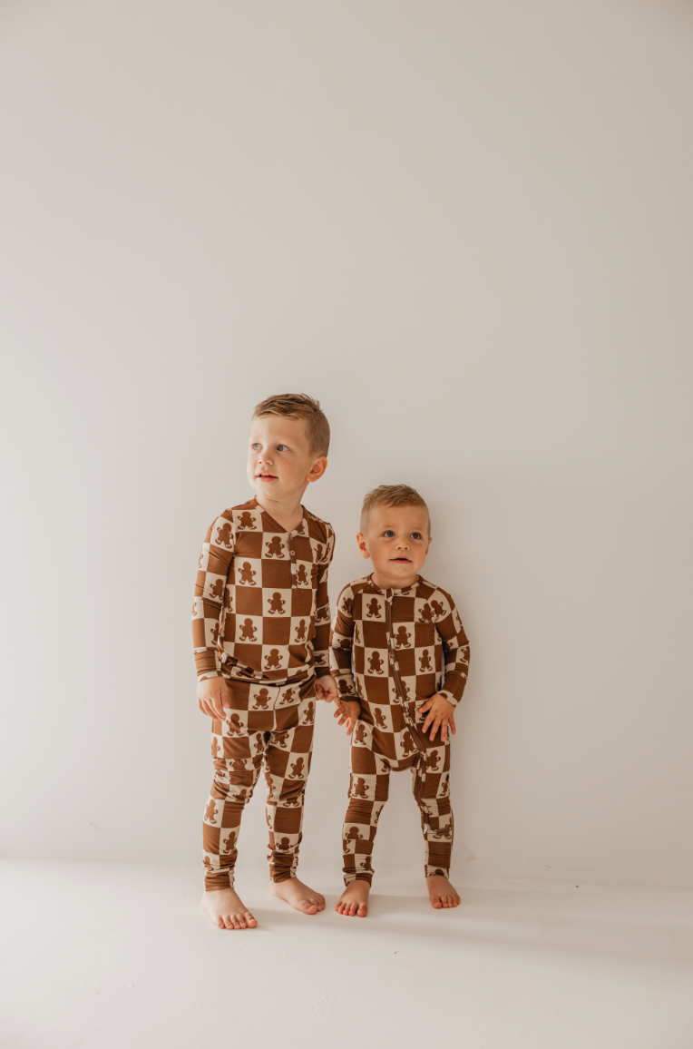 Two young children stand barefoot against a plain white wall, wearing matching forever french baby Bamboo Zip Pajamas in the Gingerbread Checkerboard design. The pajamas are crafted from breathable, hypo-allergenic bamboo fabric and feature a checkered pattern with bear prints. The older child looks to the side while the younger gazes forward.