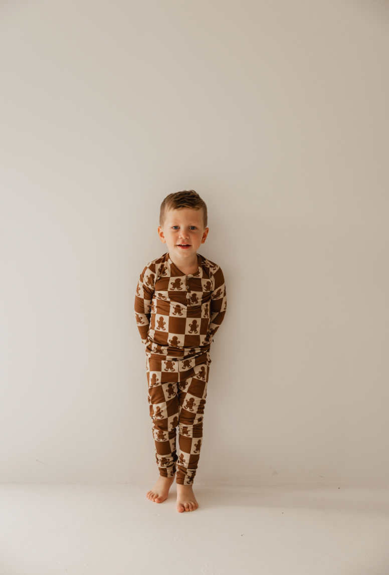 A young boy stands against a plain white wall, wearing the "Bamboo Two Piece Pajamas | Gingerbread Checkerboard" by forever french baby. The brown and white checked pajama set features charming teddy bear patterns and is crafted from breathable bamboo fabric, perfectly complementing his neutral expression. He is barefoot on a smooth surface with his arms relaxed by his sides.