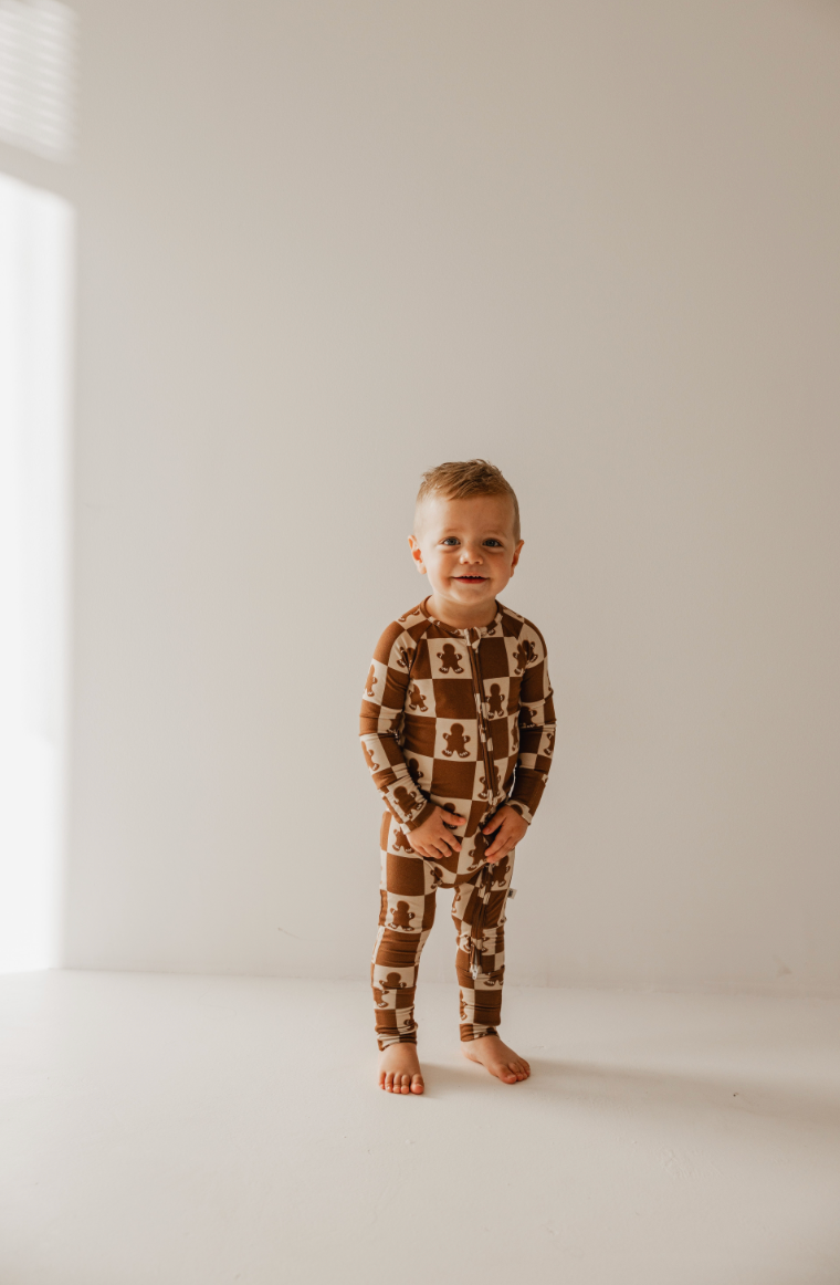 A toddler wearing hypo-allergenic Bamboo Zip Pajamas in the Gingerbread Checkerboard pattern from forever french baby stands barefoot on a light-colored floor against a plain wall. Soft sunlight filters in from the left, casting gentle shadows.