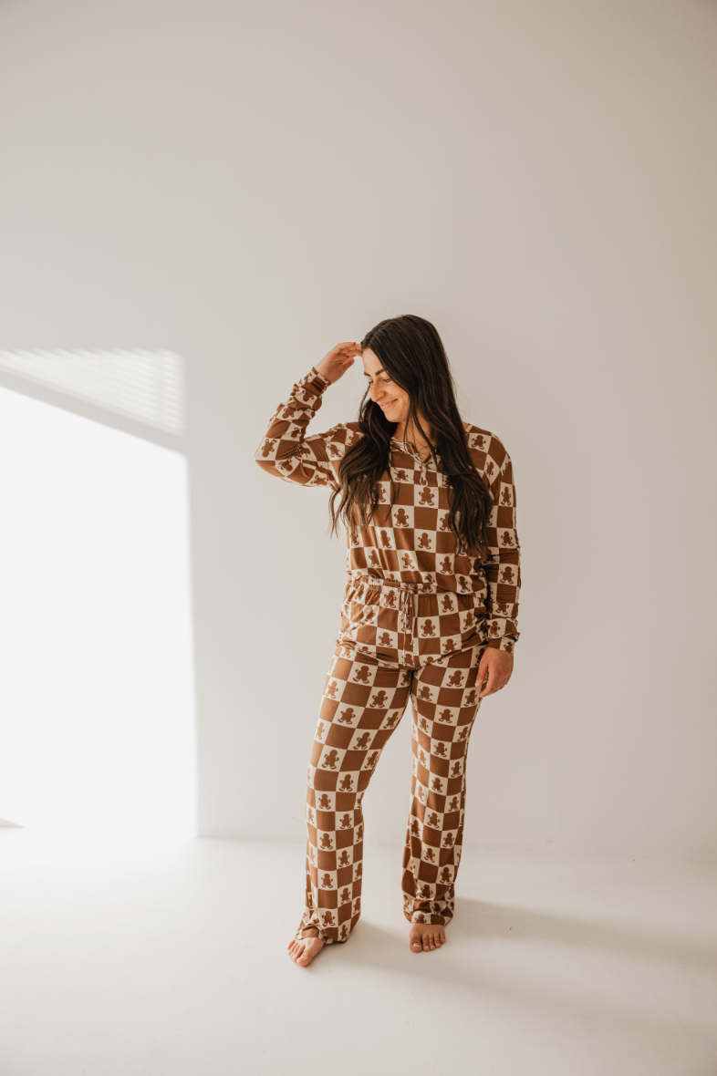 In a well-lit room, a person with long dark hair stands smiling brightly against a plain white background. They are wearing charming Women's Bamboo Pajamas in a Gingerbread Checkerboard pattern from forever french baby. The stylish rollover cuffs perfectly complement their beaming expression as they gently touch their hair while looking down.