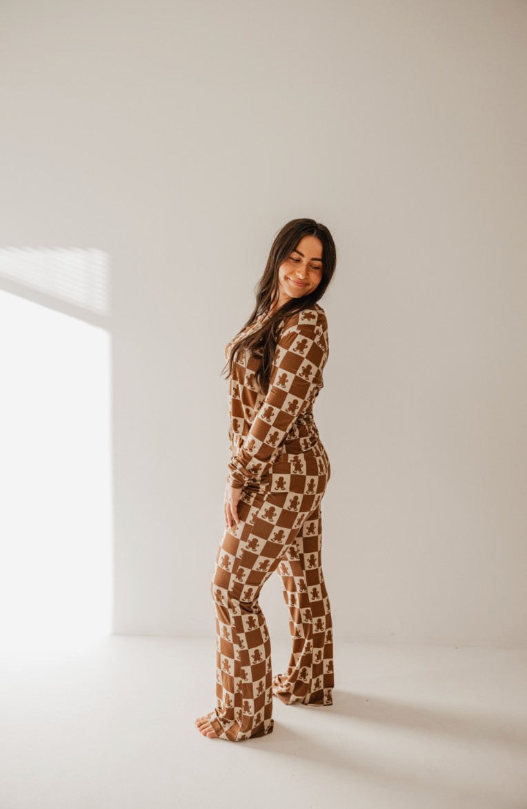 A woman with long dark hair smiles while standing barefoot in the Women's Bamboo Pajamas in the Gingerbread Checkerboard pattern, featuring rollover cuffs. She is against a plain white background, with sunlight streaming in from the left. This stylish outfit is by forever french baby.