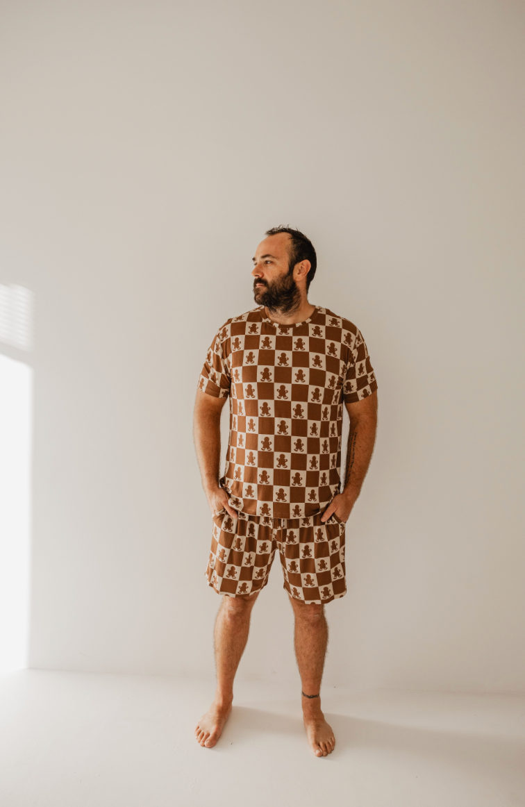 A bearded man stands barefoot against a plain backdrop, dressed in the Gingerbread Checkerboard Short Sleeve Men's Bamboo Pajamas from forever french baby. The lighting casts a gentle shadow on the wall, adding an element of tranquility to the scene.