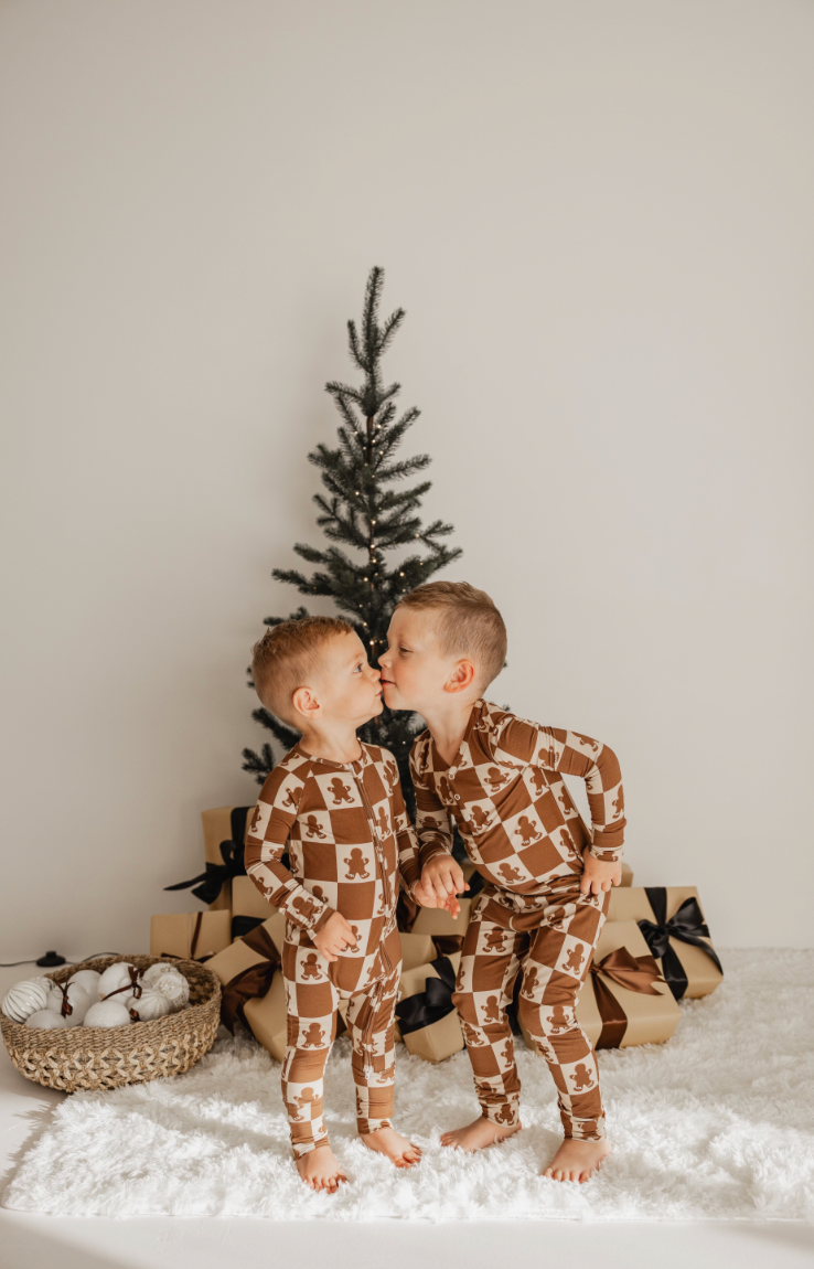 Two young children wearing matching Bamboo Two Piece Pajamas in the Gingerbread Checkerboard pattern from forever french baby stand on a white fluffy rug, sharing a kiss. A small Christmas tree with lights and wrapped gifts glows warmly in the background, adding to the festive atmosphere.