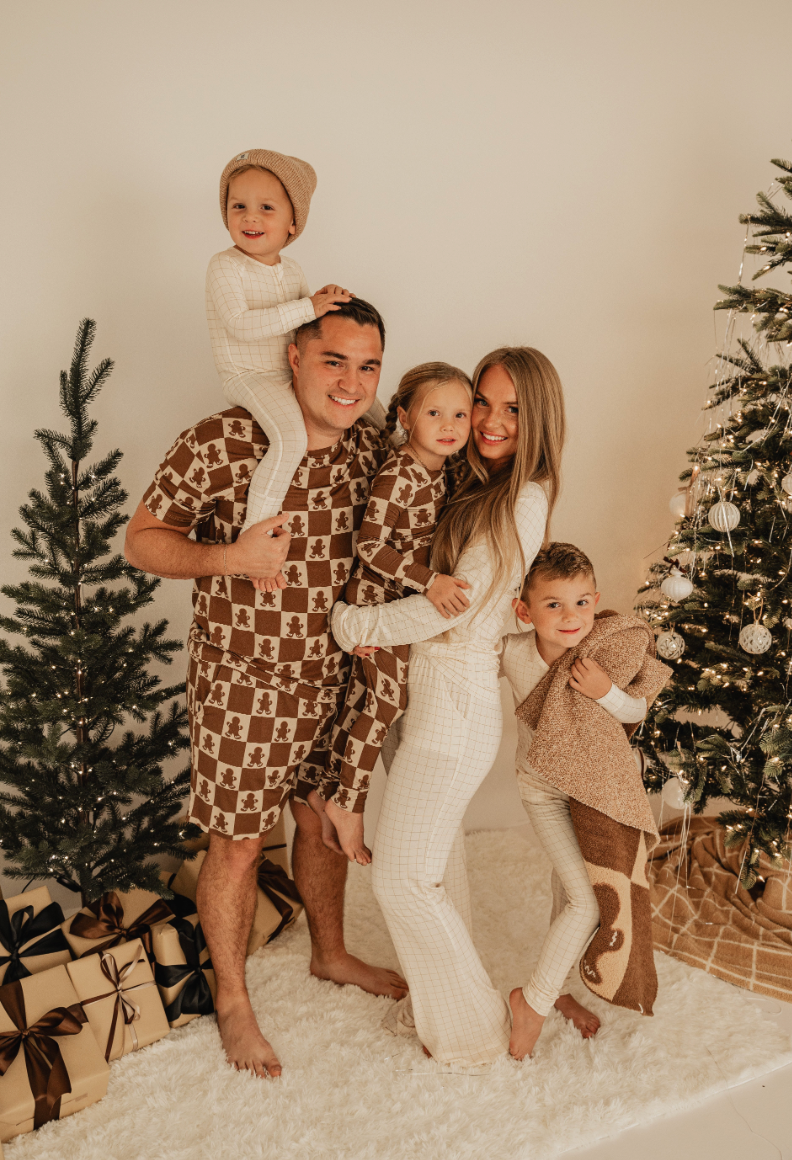 A smiling family in matching, breathable outfits poses by decorated Christmas trees and wrapped gifts. The parents hold their two young children while the older child stands beside them on a fluffy white rug, all dressed in hypoallergenic clothing from the forever french baby collection featuring Bamboo Two Piece Pajamas in Gingerbread Checkerboard.