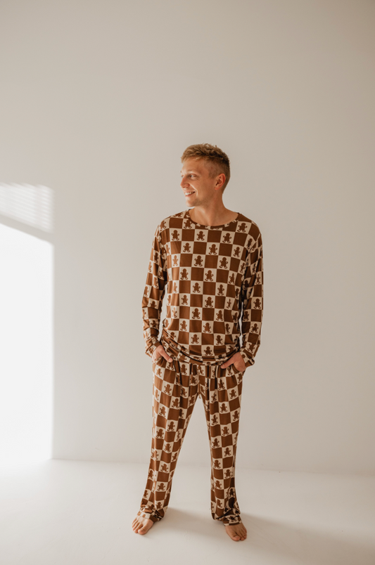 A person stands barefoot in a sunlit room, smiling and looking to the side. They are wearing cozy, matching brown and white checkered pajamas with a teddy bear pattern from Forever French Baby's Men's Bamboo Pajamas collection in Gingerbread Checkerboard. The plain background highlights the relaxed and cozy atmosphere.