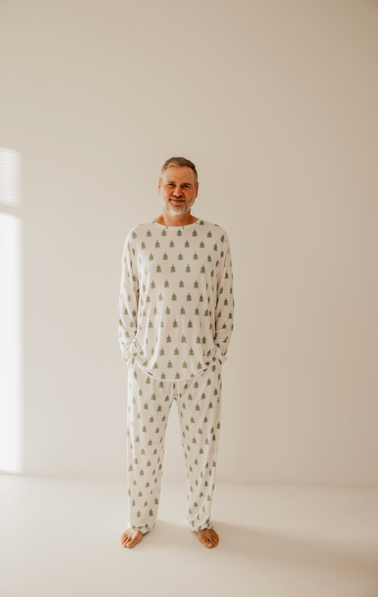 A man stands barefoot in a minimal room, wearing cozy "O Christmas Tree" bamboo pajamas by forever french baby, featuring small green tree patterns, softly lit from the left.