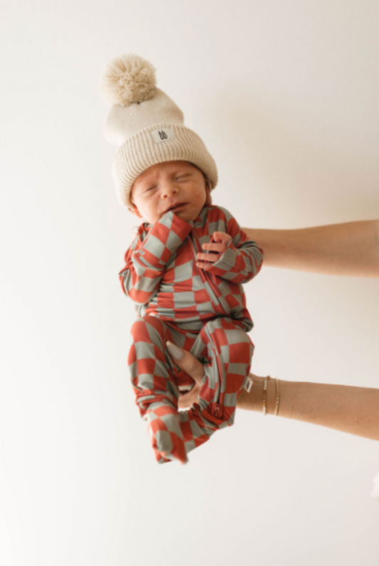 A sleeping newborn in a cream knitted hat with a pom-pom is cradled on a plain white background, dressed in Forever French Baby's breathable Retro Christmas Bamboo Zip Pajamas with red and green checks.