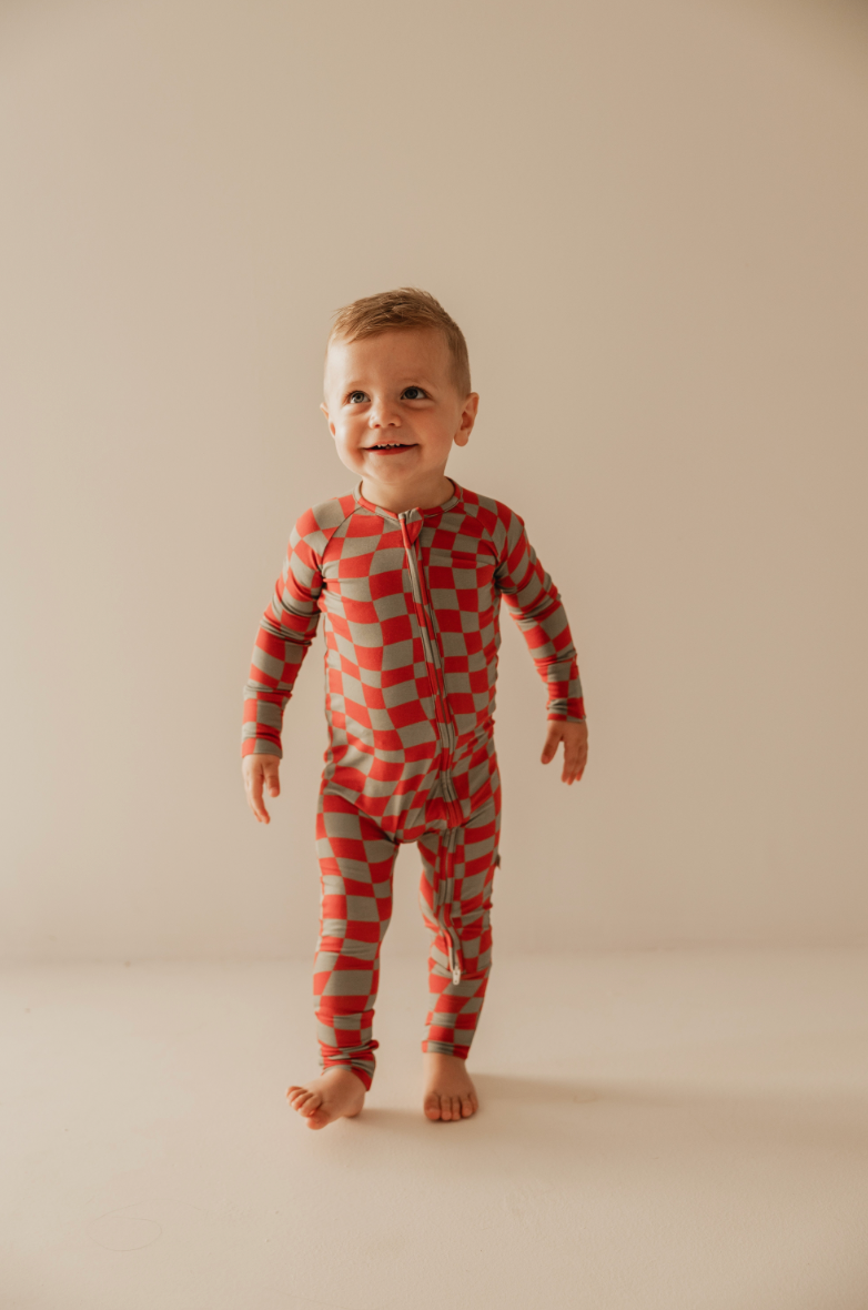 A smiling young child in a "Retro Christmas" bamboo zip pajama by forever french baby stands barefoot on a light-colored floor against a plain background.