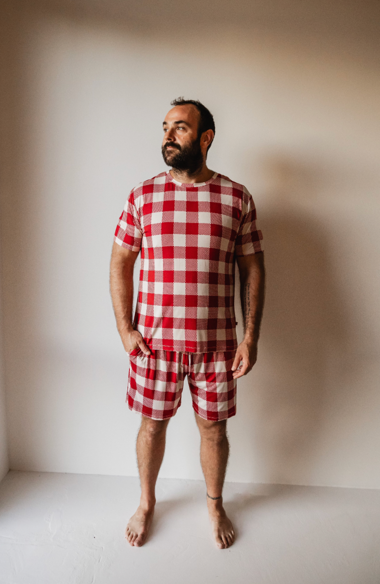 A man stands barefoot against a white wall, wearing Classic Christmas Adult Bamboo Short Pajamas by forever french baby. He gazes thoughtfully to the side, as if dreaming of cozy nights in premium bamboo sleepwear.