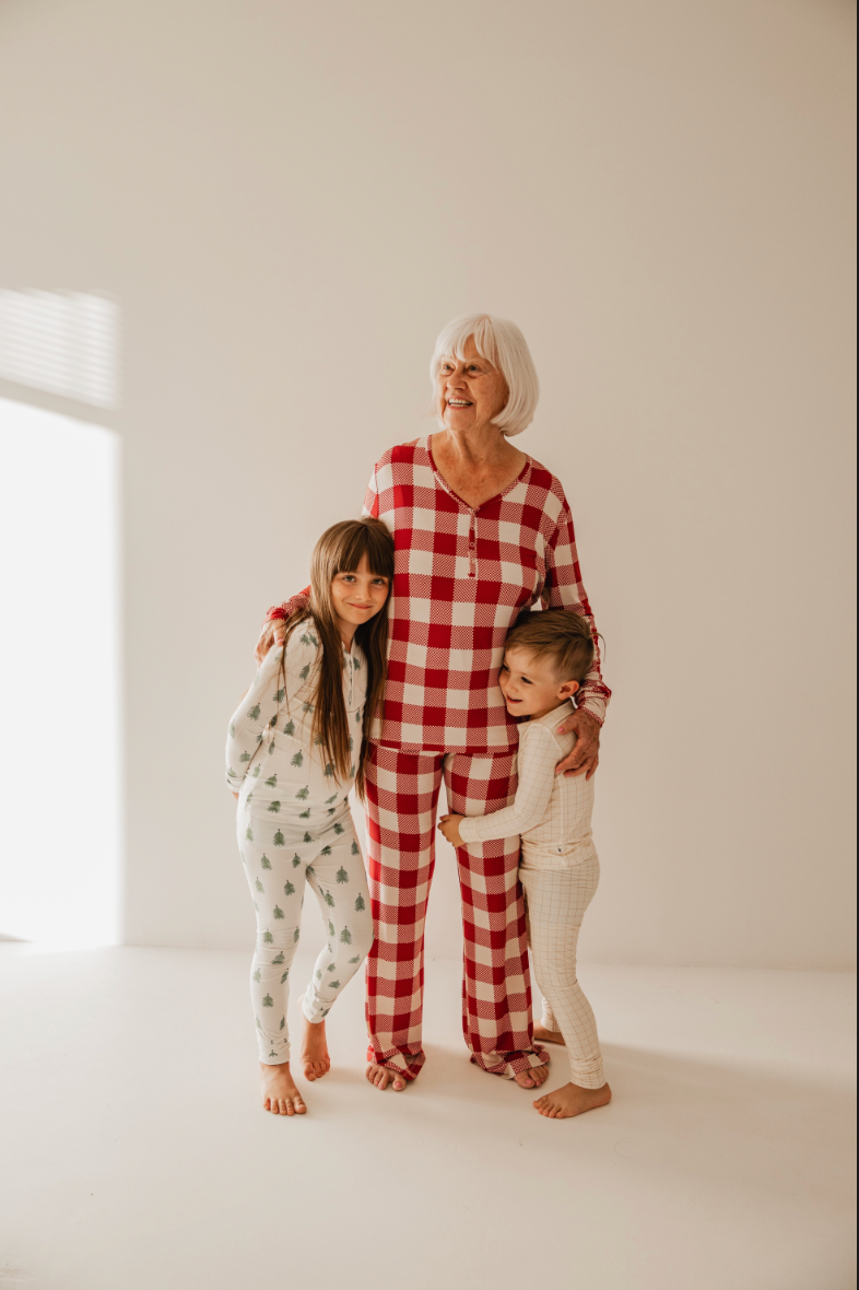 An elderly woman with white hair smiles in her red and white checkered pajamas (Classic Christmas, forever french baby). She embraces two children—one in tree-print bamboo pajamas, the other in plaid. Soft light highlights the cozy ambiance from Forever French Baby.