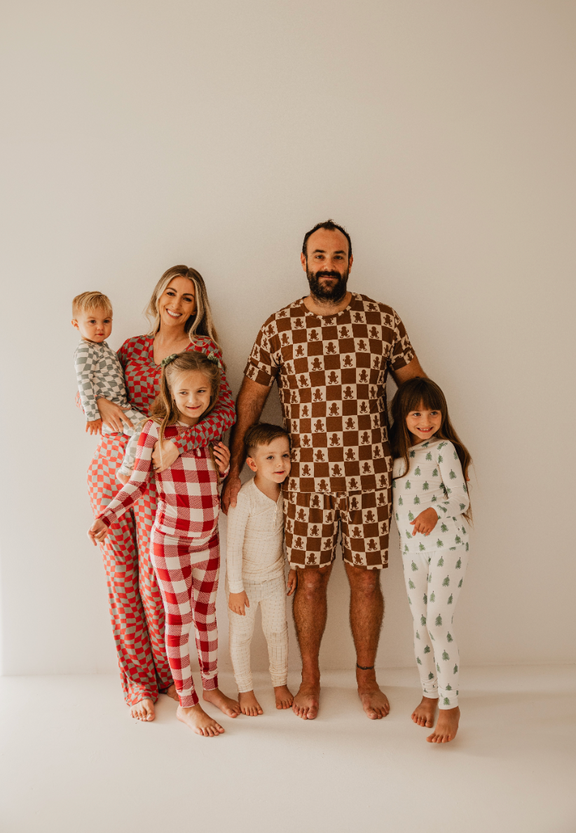 A family of six poses against a plain white background, all dressed in cozy, colorful patterned pajamas. The parents hold a toddler while the three children stand beside them, grinning in their Gingerbread Checkerboard premium bamboo pajamas—ideal for cherished moments with Forever French Baby style.