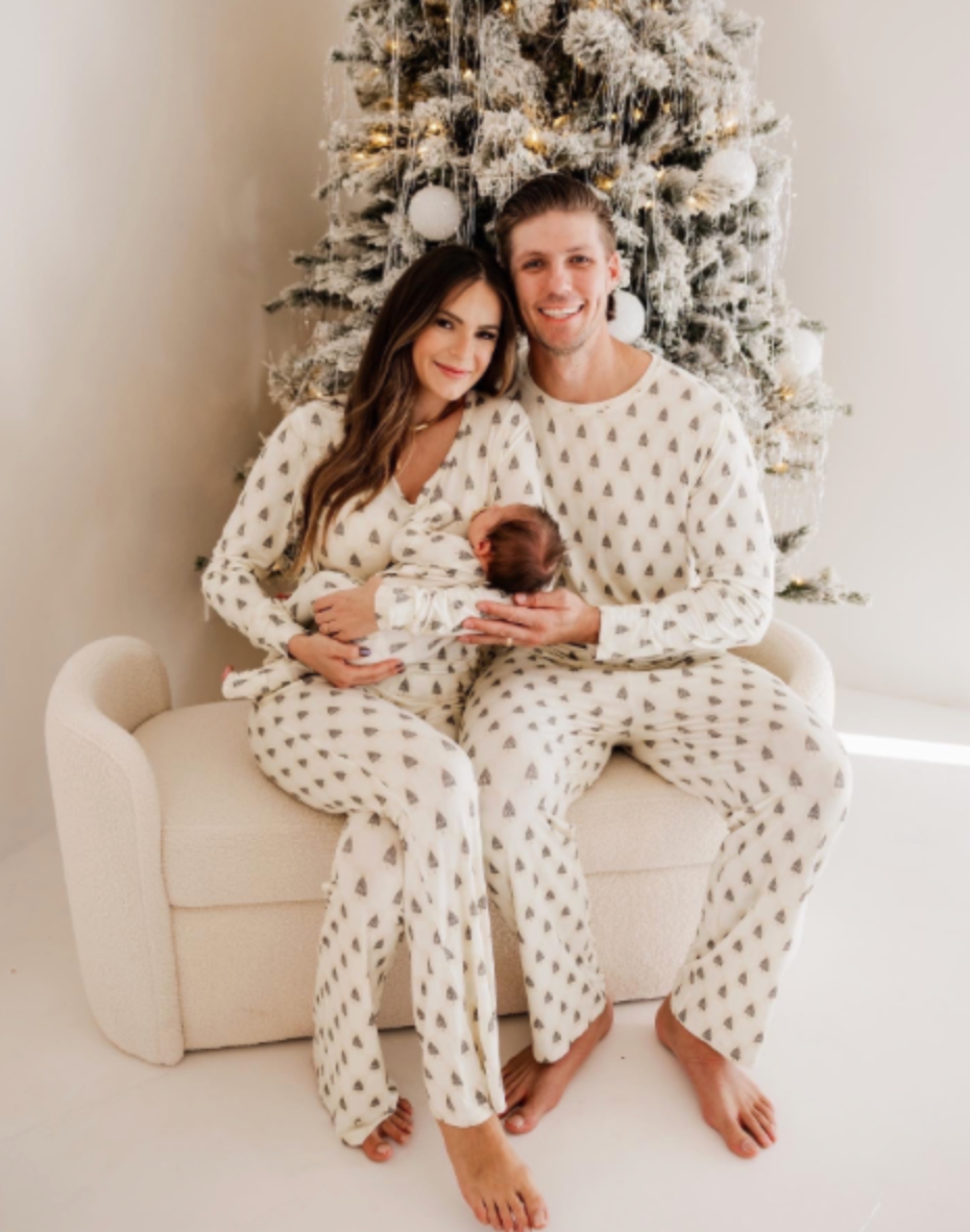 A couple in matching Vintage Christmas Deck the Halls Checkerboard pajamas by Forever French Baby sits on a snug chair in front of a white Christmas tree with silver and gold ornaments, savoring a cozy holiday moment together.