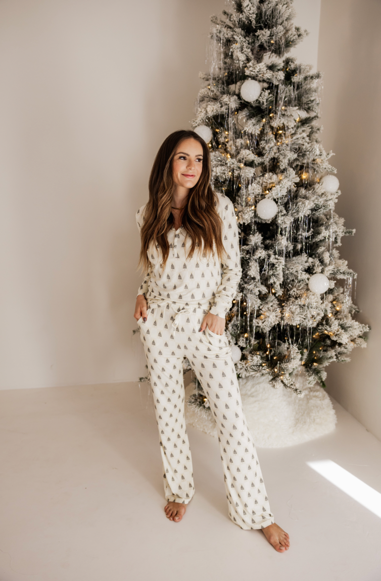 A woman poses barefoot in Forever French Baby's Vintage Christmas Deck the Halls Checkerboard bamboo pajamas beside a minimally decorated Christmas tree with white and gold ornaments, evoking a cozy atmosphere.