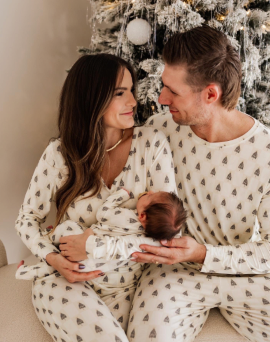 A couple in matching "Vintage Christmas Deck the Halls Checkerboard" bamboo pajamas from forever french baby sits by an elegantly adorned tree, smiling at each other. The woman holds a baby, adding to the cozy ambiance of their premium bamboo attire.