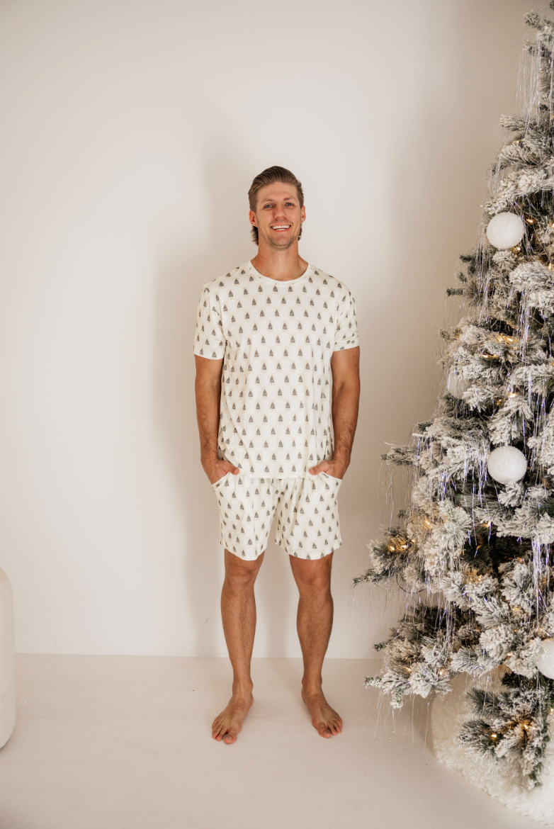 Wearing Forever French Baby's Vintage Christmas Deck the Halls Checkerboard adult bamboo short pajamas with rollover cuffs, a person smiles next to a Christmas tree adorned in white and gold ornaments, creating a cozy holiday atmosphere. Perfect for those Forever French Baby moments.