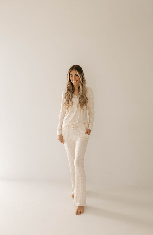 A woman with long, wavy hair stands barefoot against a plain background, wearing a matching long-sleeve top and pants set in a soft, light-colored checkered pattern. Made of premium bamboo, this ensemble is named Women's Bamboo Pajamas | Golden Grid. She smiles warmly with her hands in her pockets, perfectly embodying the essence of cozy sleepwear from forever french baby.