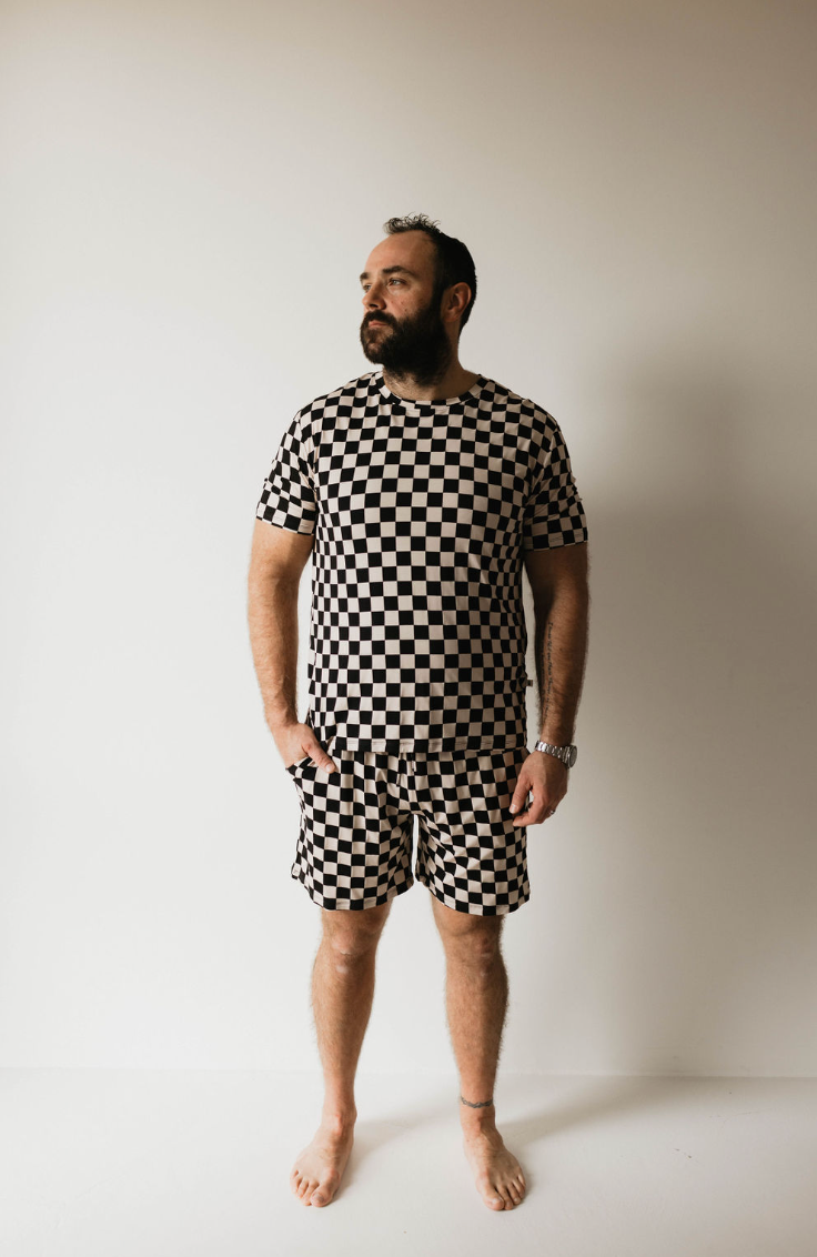 A man stands barefoot against a plain white background, wearing the forever french baby Adult Bamboo Short Pajamas in Black Checkerboard. He is looking off to the side, with his hands relaxed by his sides. He has a beard and a watch on his left wrist.