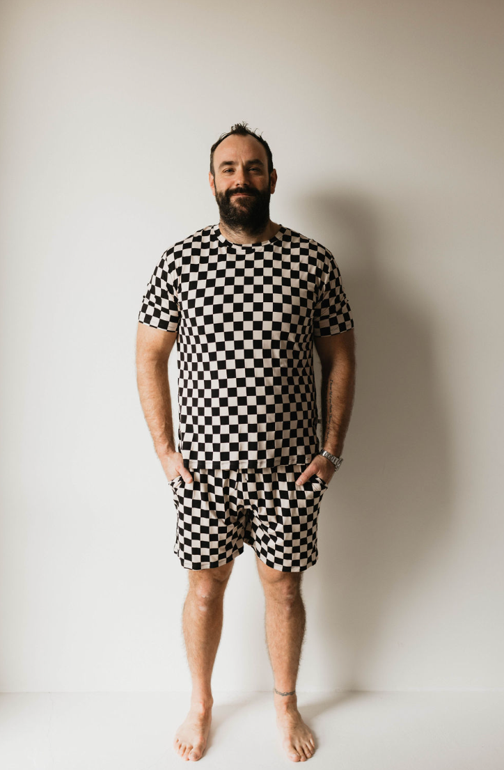 A bearded man stands barefoot against a plain white background. Clad in "Adult Bamboo Short Pajamas | Black Checkerboard" from forever french baby, he exudes a laid-back vibe. His arms are relaxed at his sides with hands in his pockets, showcasing a watch on his left wrist and an anklet on his right ankle.