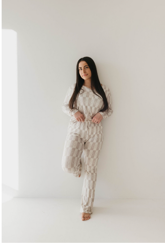 A person with long dark hair stands against a white wall, wearing forever french baby's Women’s Bamboo Pajamas in the Smile Checkerboard pattern. They have a relaxed expression, with one leg slightly bent and hands resting on the drawstring waist of their hypo-allergenic loungewear.