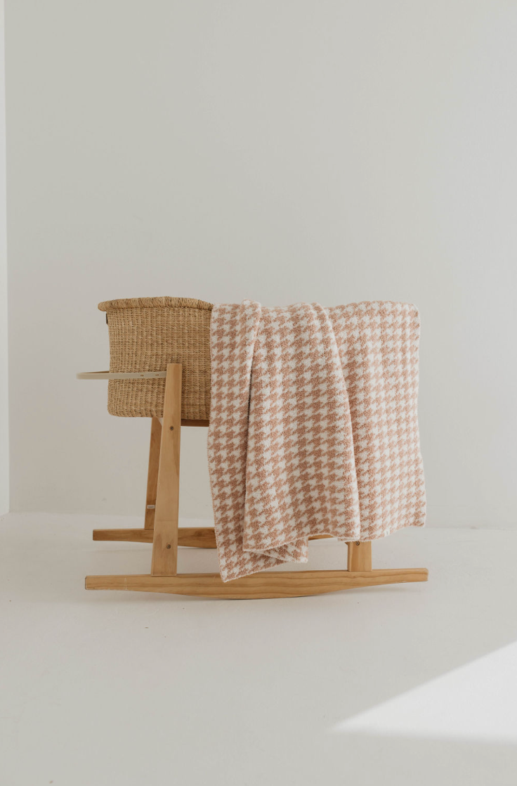 A wooden rocking cradle with a wicker basket is placed in a well-lit, minimalistic room. A Plush Blanket | Houndstooth Rosé & Cream by forever french baby is draped over one side of the cradle. The background is plain white.