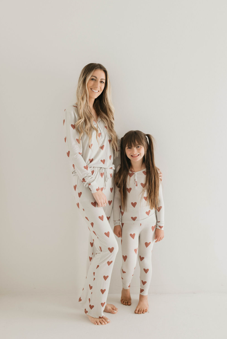 A woman and a young girl stand side by side against a white background, both smiling and wearing matching Women's Bamboo Pajamas from forever french baby, adorned with Queen Of Hearts Paisley’s Hand Drawn Print. The woman has long blonde hair, while the girl sports long brown hair with bangs and pigtails. Both are barefoot.