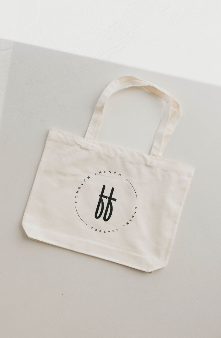 A Forever French | Canvas Tote from forever french baby, with two handles, lies flat on a grey and white surface. The durable white canvas bag features a circular logo with the letters "FF" in the center and the words "FOREVER FRENCH" repeated around the circle.