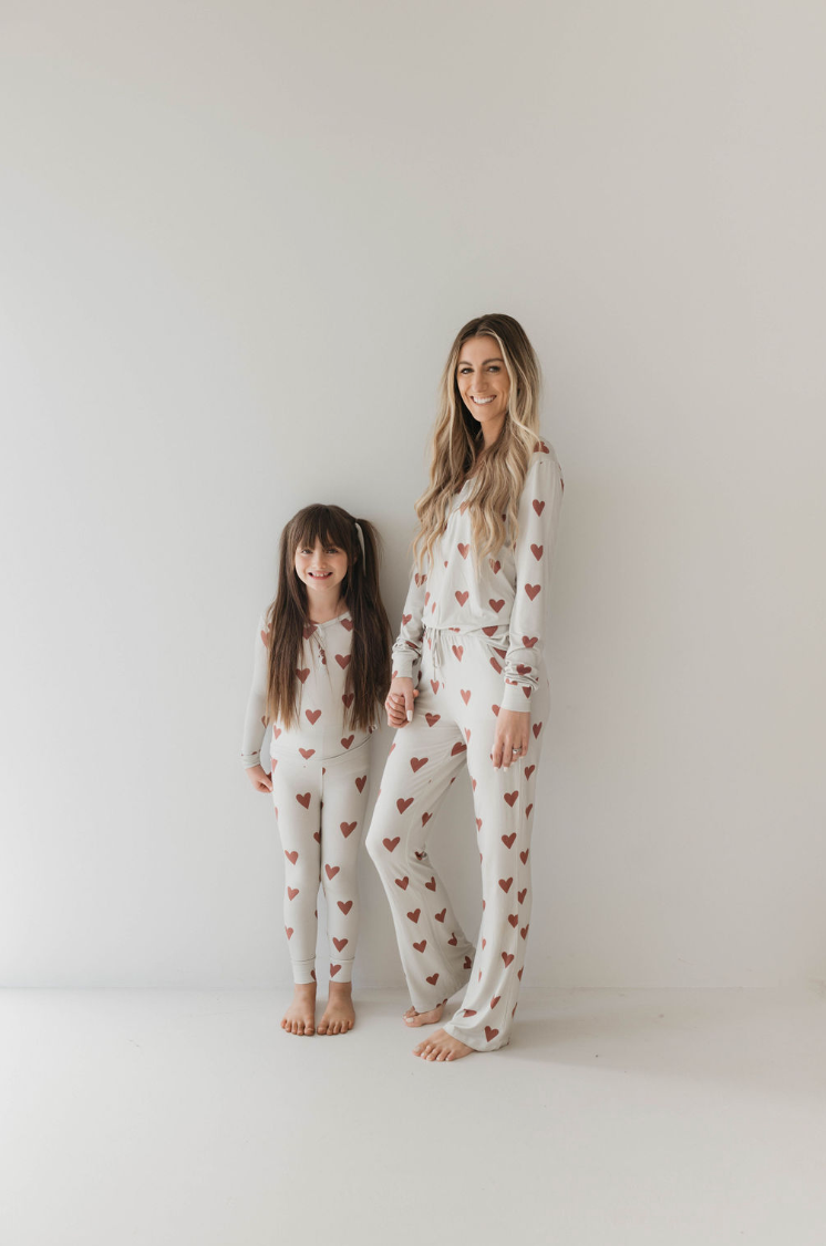 A woman and a young girl stand against a plain white background, holding hands and wearing Women's Bamboo Pajamas | Queen Of Hearts Paisley’s Hand Drawn Print by forever french baby. Both are barefoot, have long hair, and the woman smiles at the camera.