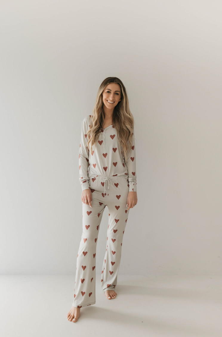 A woman smiles while wearing the Women's Bamboo Pajamas from forever french baby, featuring the Queen Of Hearts Paisley’s Hand Drawn Print. She stands barefoot on a plain light-colored floor against a simple off-white wall, looking every bit like royalty in her cozy apparel.