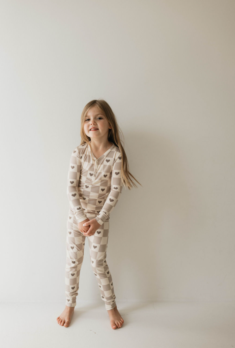 A young girl with long hair is standing against a plain white background, smiling. She is wearing the "Bamboo Two Piece Pajama | Check Your Heart" from forever french baby, featuring beige and white checkered patterns adorned with heart designs. The footless pajamas add to her playful expression as she stands barefoot and cheerful.