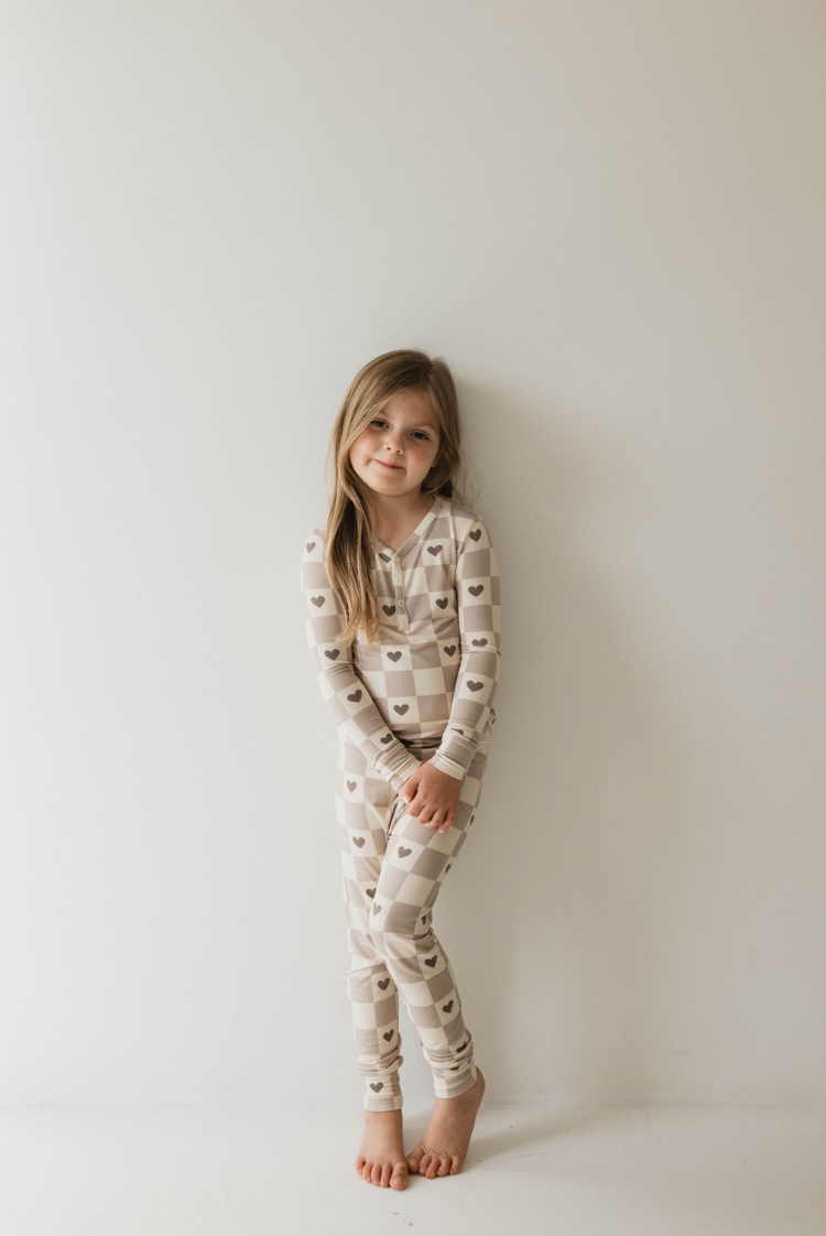 A young girl stands against a white wall, smiling gently. She is dressed in the cozy "Check Your Heart" Bamboo Two Piece Pajama by forever french baby, featuring a beige and white checkered pattern with small brown hearts. She has long, light brown hair and is barefoot in an overall minimalistic and bright setting.