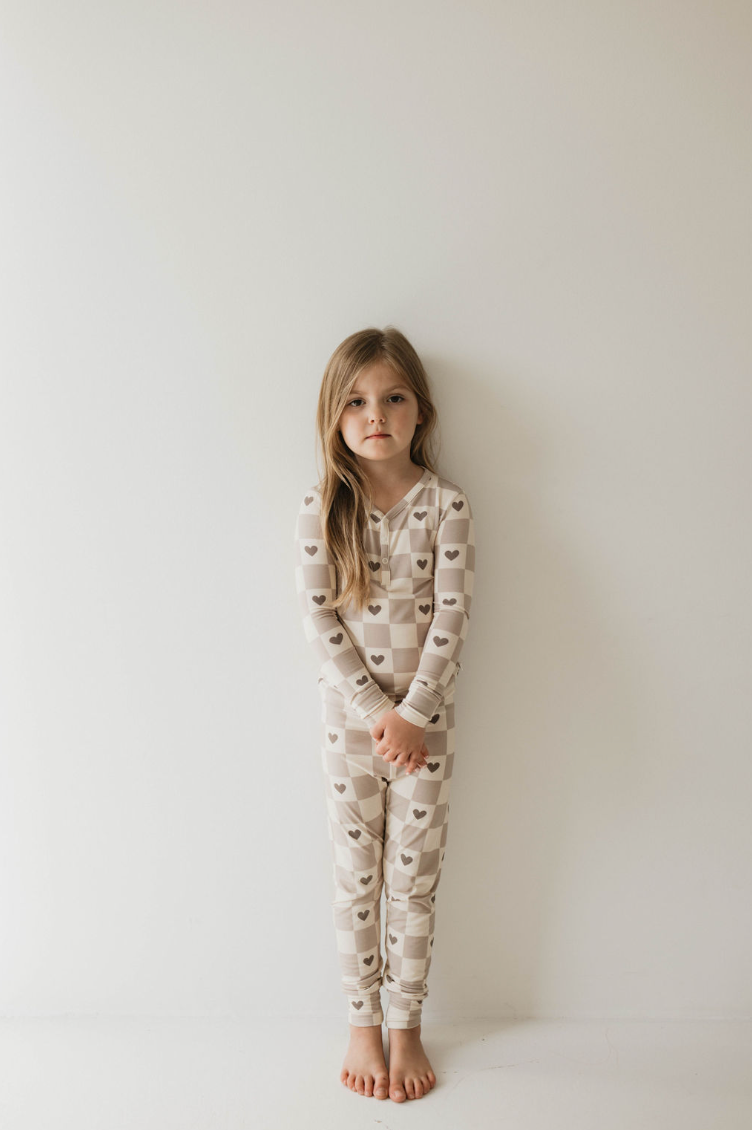 A young child with long, brown hair is standing against a plain white wall. They are wearing the forever french baby Bamboo Two Piece Pajama from the Check Your Heart collection, featuring a beige and white checkered pattern adorned with brown heart shapes. The child is barefoot and has a slight, serious expression on their face, arms relaxed at their sides.
