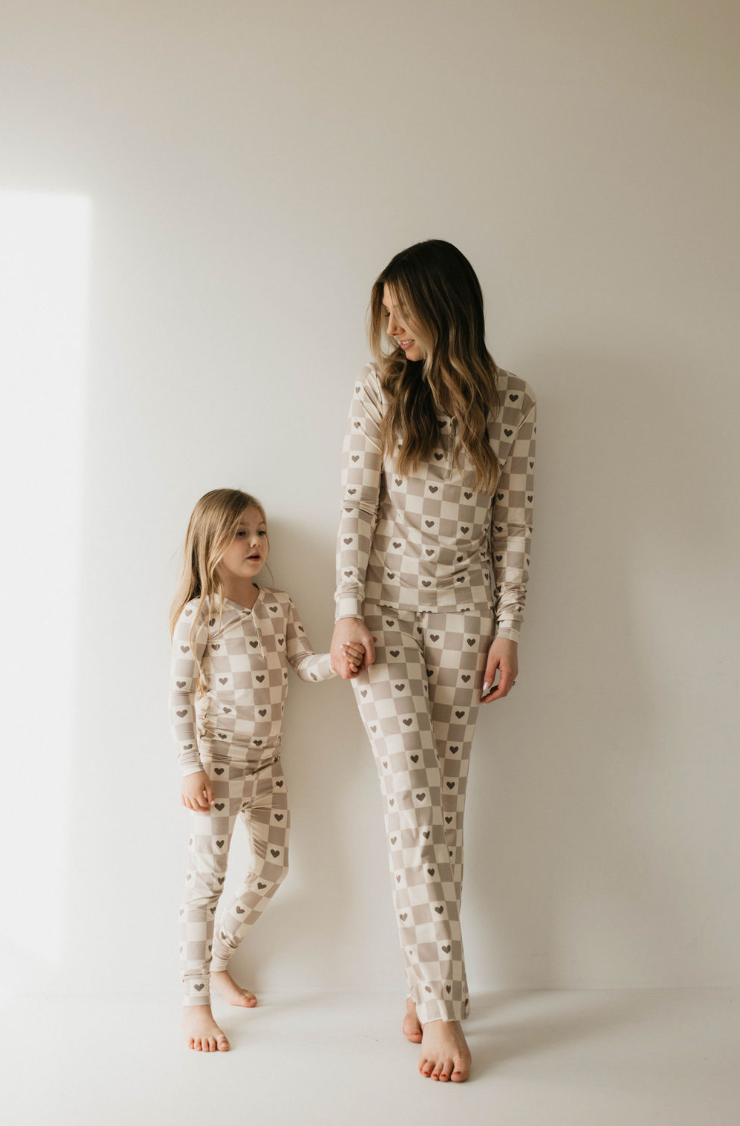 An adult and child stand against a white wall, holding hands and gazing at each other. They are both clad in matching Bamboo Two Piece Pajamas from forever french baby, featuring a checkered heart pattern. The child is barefoot, and a sliver of sunlight casts light on part of the wall.