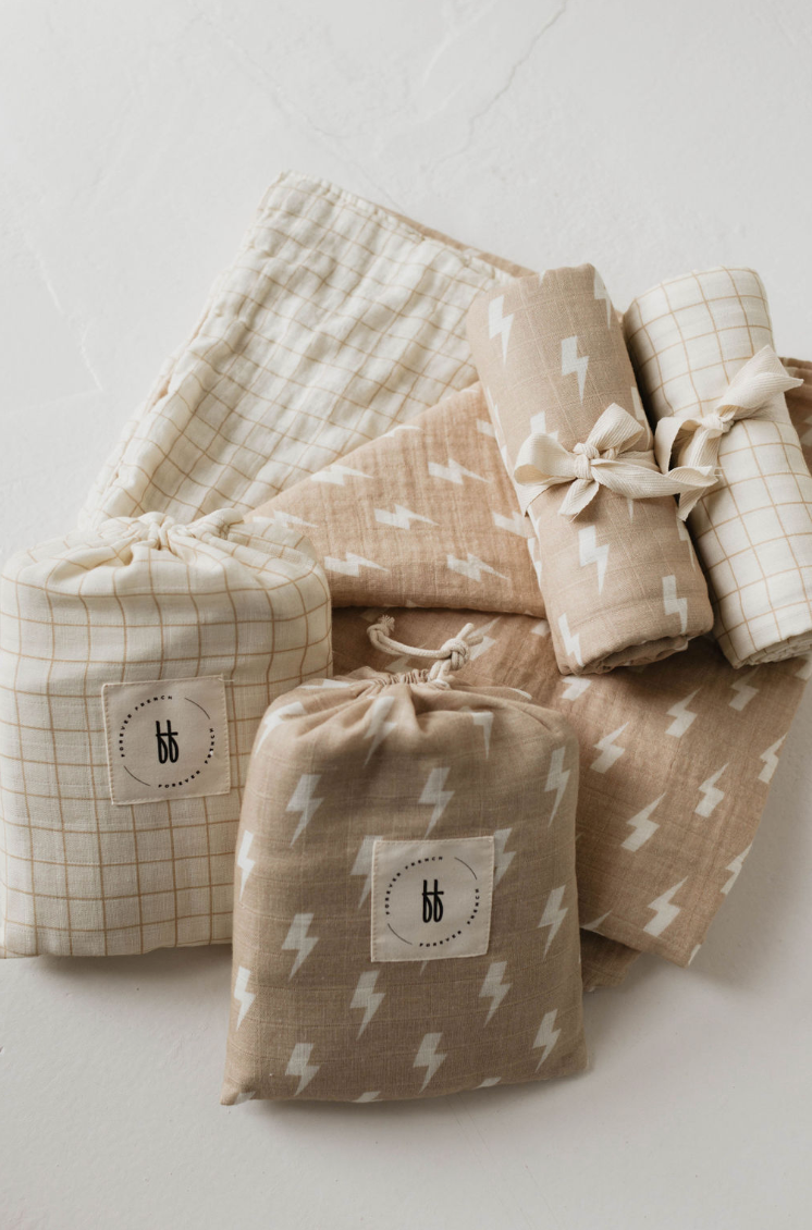 A collection of Muslin Quilts by forever french baby in tan and cream lightning bolt and golden grid patterns lies on a white surface. Two drawstring bags, each made from 6 layers of muslin cotton, feature a logo with the initials "bb" on a circular label.