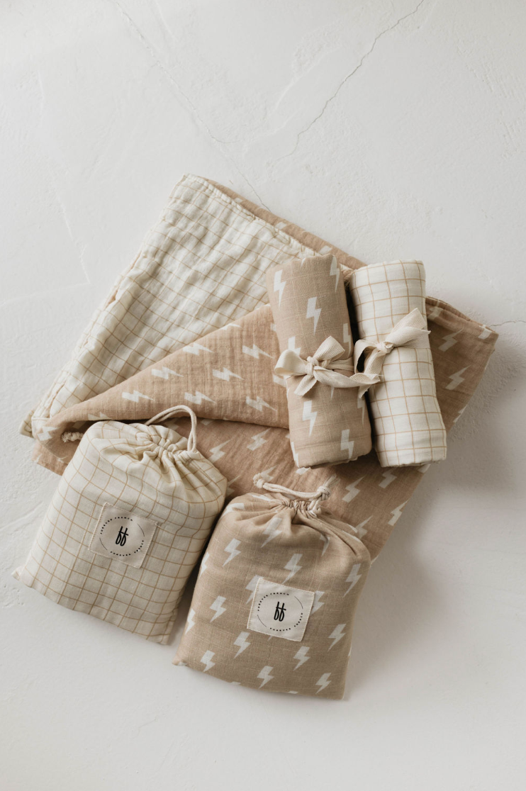 A neatly arranged display of baby essentials on a white surface. Items include a light beige blanket with a checkered pattern, another 6 layers of the Muslin Quilt featuring white lightning bolts and golden grid from Forever French Baby, and two drawstring pouches with patterned fabric. Everything exudes the timeless charm of Forever French Baby.