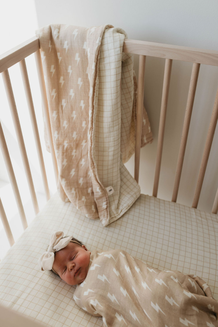 A newborn baby sleeps peacefully in a crib, swaddled in the Tan and Cream Lightning Bolt & Golden Grid Muslin Quilt from forever french baby. A similar quilt and a cream-colored blanket with a grid pattern hang over the side of the crib. The soft, 6 layers of muslin cotton ensure comfort. The baby wears a matching headband from Forever French Baby.