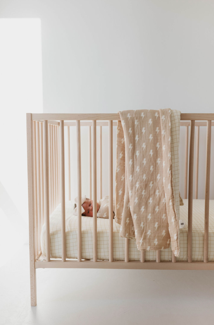 Image of a minimalist baby crib made of light wood placed in a bright room. The crib features a beige and white checkered mattress, complemented by the Tan and Cream Lightning Bolt & Golden Grid Muslin Quilt from Forever French Baby draped over the side. A small plush toy is partially visible inside the crib.