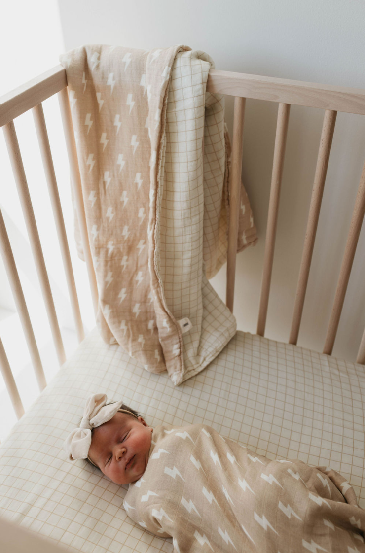 A sleeping baby is swaddled in a beige blanket with white lightning bolt patterns, lying in a light wooden crib. Two blankets, including a Tan and Cream Lightning Bolt & Golden Grid reversible muslin quilt from forever french baby, are draped over the crib's side. The baby has a bow headband.