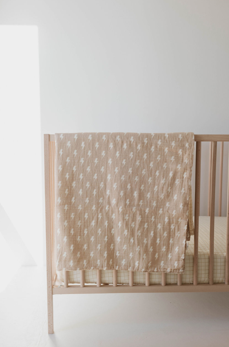 A light wood baby crib sits in a bright, minimalist room. A Tan and Cream Lightning Bolt & Golden Grid Muslin Quilt by forever french baby, crafted with six layers of muslin cotton, hangs neatly over the crib's side. The crib mattress features a light, checkered sheet. Sunlight streams in from the left, casting a soft glow on the scene.