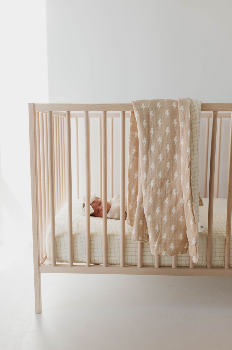 A baby lies in a light wooden crib with a beige mattress. Draped over the side of the crib is a Tan and Cream Lightning Bolt & Golden Grid reversible muslin quilt by Forever French Baby, made from 6 layers of muslin cotton. The bright room features minimalist decor, capturing the timeless elegance associated with Forever French Baby.