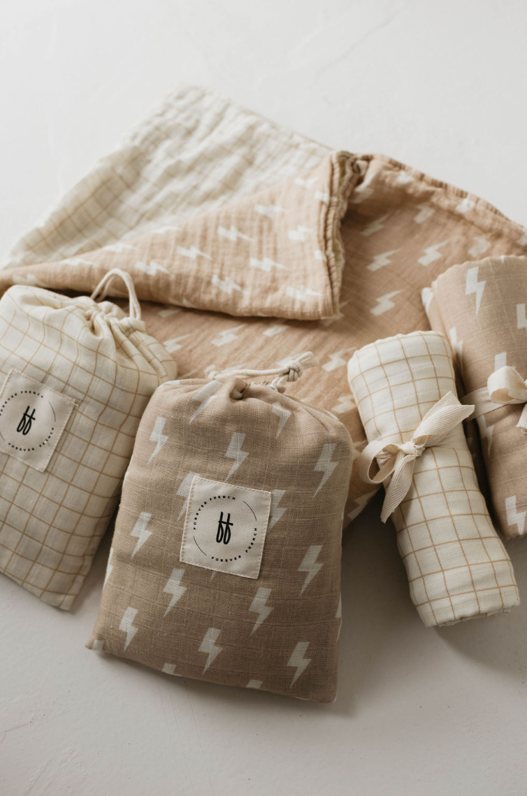 A collection of soft fabric items in neutral tones lies on a white surface. The items include Forever French Baby pouches with drawstrings, a folded Muslin Swaddle in Tan & Cream Lightning Bolt, and a tied bundle. The fabrics feature patterns of grids and lightning bolts in light beige and white, perfect for any baby's delicate needs.