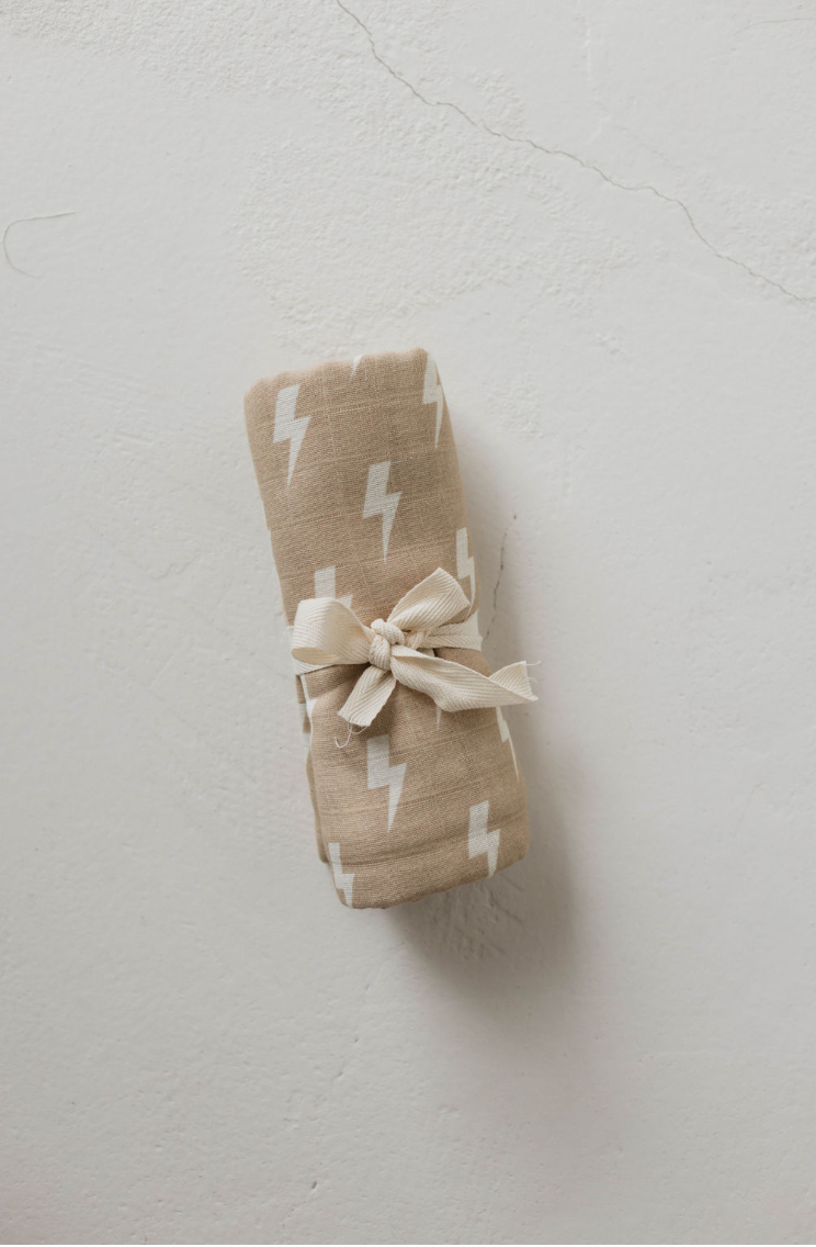 A rolled-up "Muslin Swaddle | Tan & Cream Lightning Bolt" from forever french baby, tied with a bow, is placed on a light-colored textured surface. This swaddle is part of the exclusive Forever French Baby collection.