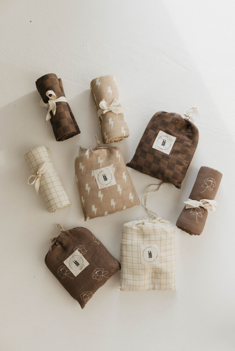 An assortment of rolled and folded fabric bundles, showcasing various brown, beige, and white patterns including checks, lightning bolts, grids, and floral prints. Each Forever French Baby muslin swaddle is tied with string or placed in matching pouches adorned with simple fabric labels for your little one. Featured product: Muslin Swaddle | Tan & Cream Lightning Bolt by forever french baby.